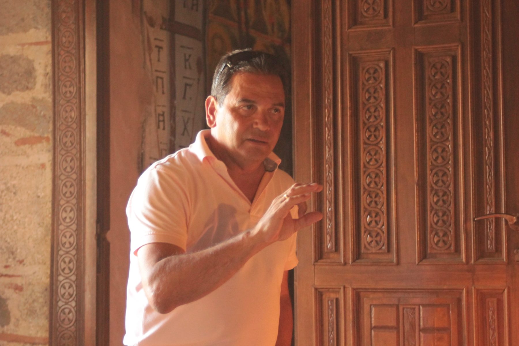 Petros mid-explanation beside a carved wooden monastery door, gesturing as he speaks.