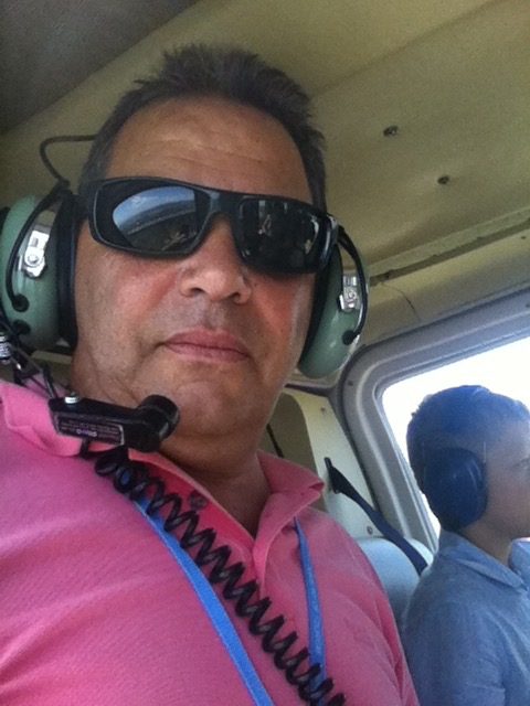 Petros in a helicopter cockpit wearing an aviation headset, with a passenger seated behind him.