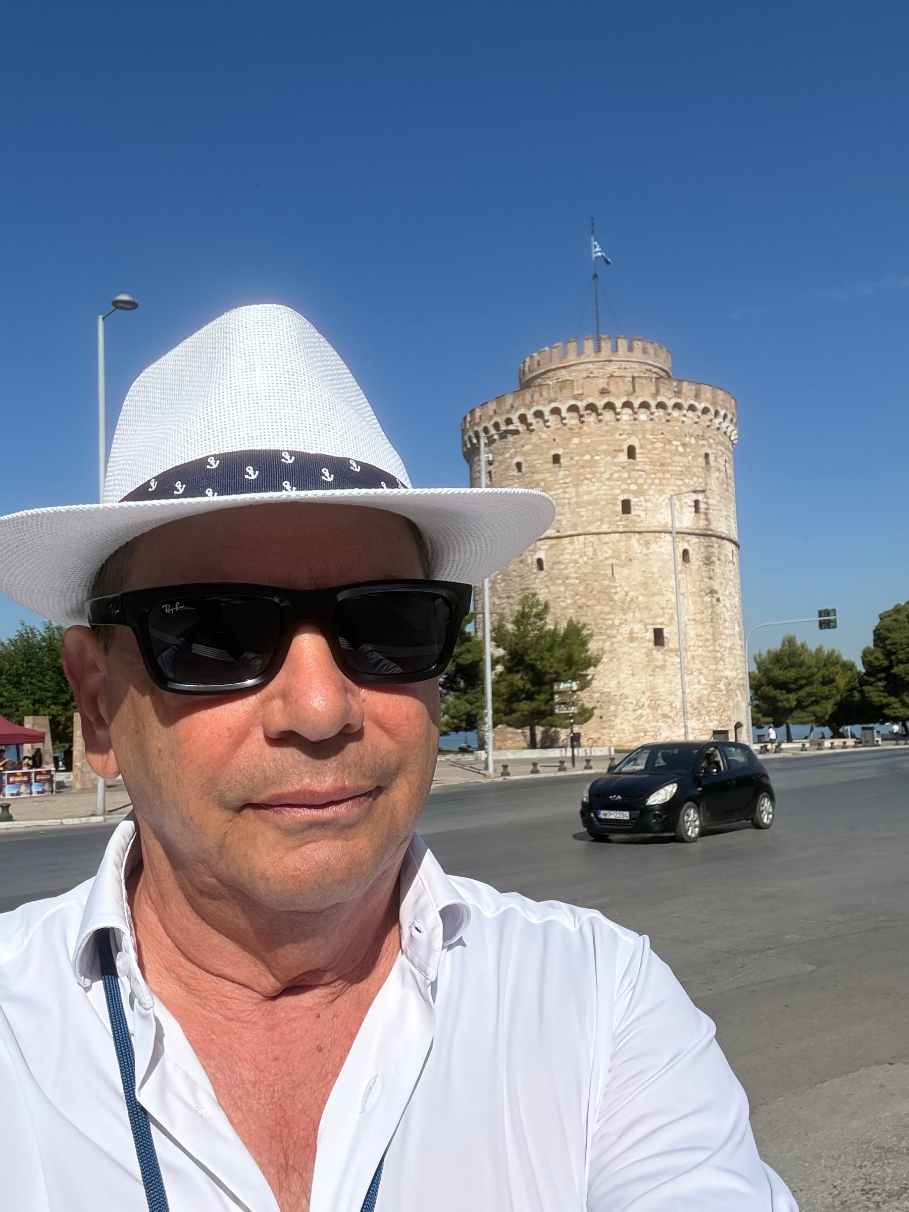 Petros Vassiliadis in a white hat and sunglasses, the White Tower of Thessaloniki behind him.