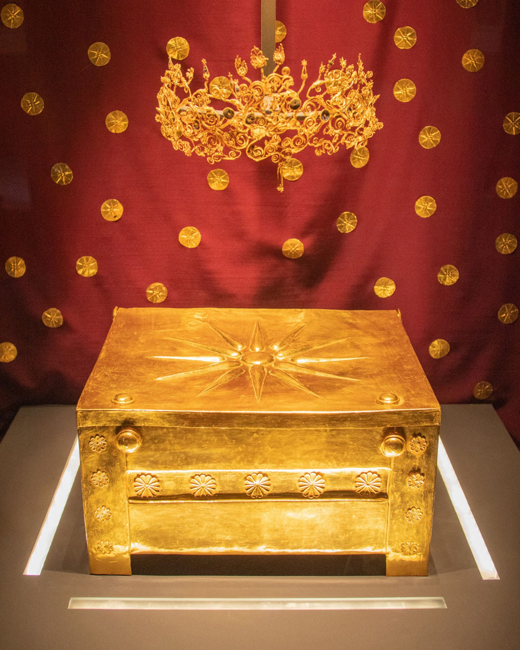 The gold larnax of Vergina, its lid bearing the Vergina Sun, with a gold wreath displayed above it.